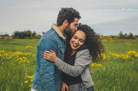 A man and woman happily embrace in a meadow, as he kisses the side of her head.